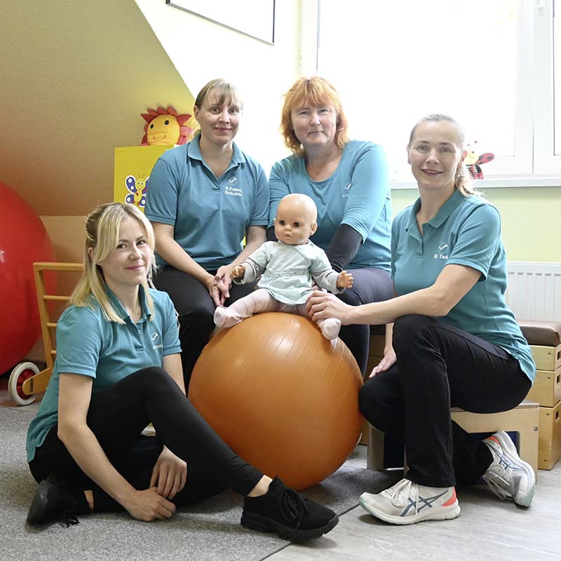 Das Therapeuten-Team für Kindertherapien der Vier Tore Reha Neubrandenburg sitzt im Behandlungsraum für ein Gruppenfoto zusammen.